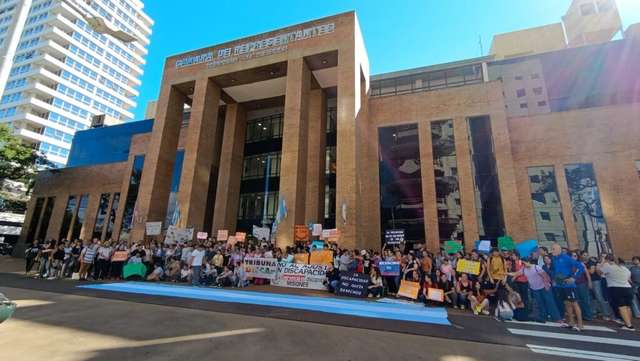 ​Una multitud se manifiesta frente al edificio de la Cámara de Representantes de Misiones. Los manifestantes sostienen pancartas con mensajes sobre discapacidad y una bandera argentina extendida en el suelo. El edificio de ladrillo visto destaca al fondo bajo un cielo despejado.
