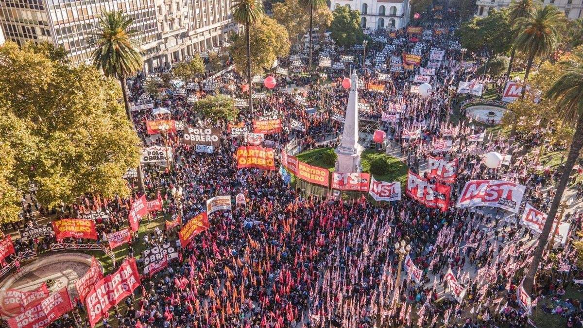 Fotografía aérea de un acto masivo del Frente de Izquierda en Plaza de Mayo, Buenos Aires. La plaza está colmada de personas con banderas rojas. Se destacan carteles del 'Frente de Izquierda de los Trabajadores' y del 'Partido Obrero'. Hay globos rojos de gran tamaño flotando sobre la multitud y edificios históricos al fondo, como la Catedral Metropolitana. Fotografía aérea de un acto masivo del Frente de Izquierda en Plaza de Mayo, Buenos Aires. La plaza está colmada de personas con banderas rojas. Se destacan carteles del 'Frente de Izquierda de los Trabajadores' y del 'Partido Obrero'. Hay globos rojos de gran tamaño flotando sobre la multitud y edificios históricos al fondo, como la Catedral Metropolitana.