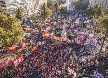 Fotografía aérea de una manifestación masiva en Plaza de Mayo, Buenos Aires. La plaza está colmada de personas con banderas rojas. Se destacan carteles del 'Frente de Izquierda de los Trabajadores' y del 'Partido Obrero'. Hay globos rojos de gran tamaño flotando sobre la multitud y edificios históricos al fondo, como la Catedral Metropolitana. Fotografía aérea de una manifestación masiva en Plaza de Mayo, Buenos Aires. La plaza está colmada de personas con banderas rojas. Se destacan carteles del 'Frente de Izquierda de los Trabajadores' y del 'Partido Obrero'. Hay globos rojos de gran tamaño flotando sobre la multitud y edificios históricos al fondo, como la Catedral Metropolitana.