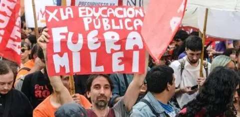 "Un manifestante sostiene en alto un cartel rojo con letras blancas que dice: 'X LA EDUCACIÓN PÚBLICA FUERA MILEI'. Se encuentra rodeado de una multitud con banderas rojas durante la marcha universitaria."