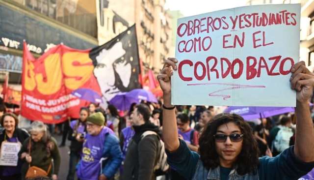 "Un joven sostiene en alto un cartel escrito a mano durante la cuarta marcha federal univeristaria. El texto en letras rojas dice: 'Obreros y estudiantes como en el Cordobazo'. Al fondo se observan banderas rojas y negras, paraguas violetas y una multitud de manifestantes."